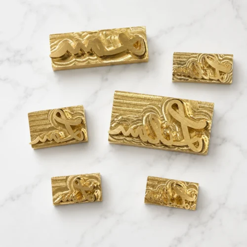 Multiple sizes of cursive script signature brass text dies arranged on white marble slab, handwritten font for custom leather name embossing.