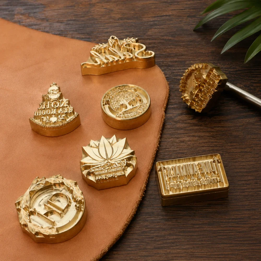 Curated lifestyle shot of lotus, tree monogram brass branding stamps with iron handle arranged on leather over dark wooden table.