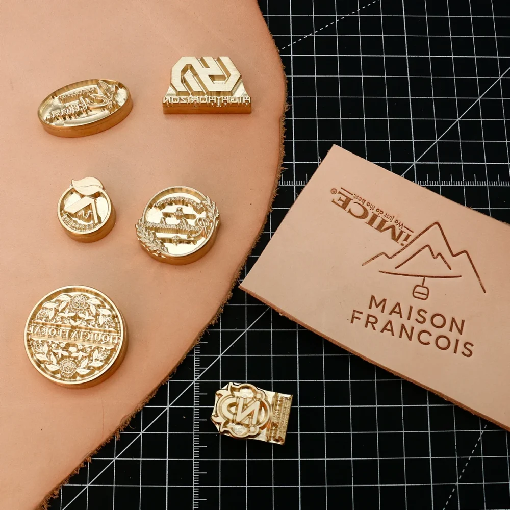 Assorted round, oval, geometric custom CNC brass logo stamps laid on leather with finished hot stamped leather sample on grid cutting mat.