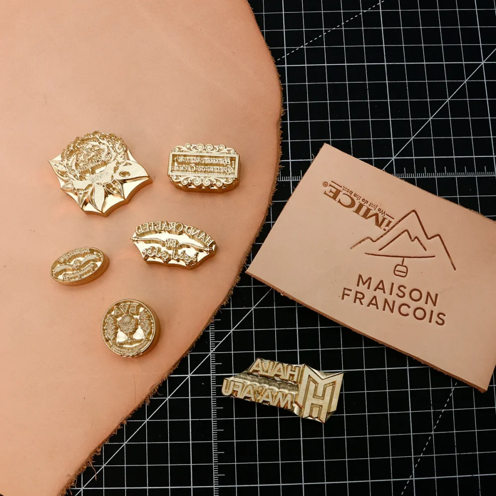 Floral badge, scroll frame, oval monogram CNC brass stamp assortment next to blind embossed leather logo test piece on grid mat.