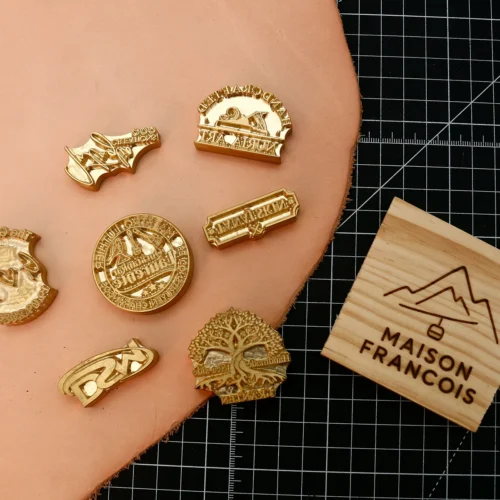 Collection of vintage script, circular seal, tree graphic CNC brass branding stamps paired with finished wood branding sample on black grid mat.